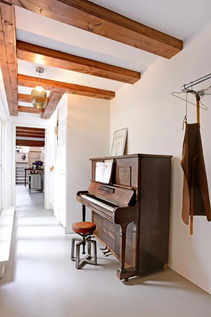 Houten piano met kruk in een lichte hal met houten plafondbalken aan de Nieuwe Kerkstraat.